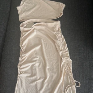Beige set - Snygg beige två delad set med omlott och snörning i midjan. Den  har en axelbandslös design med oneshoulder-topp och högt slits på sidan och tight passform.