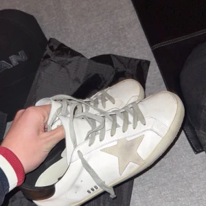 Golden Goose vita sneakers med stjärna - Säljer ett par vita Golden Goose sneakers med grå snörning och den ikoniska beige mockastjärnan på sidan. Skorna har en platt sula och svart detalj på hälen. Perfekta för dig som gillar en clean men ändå unik look. Köpte skorna för några månader sen här på plick men gillade ej stilen, kan få ner lite vid snabb affär, äkta skor såklart priset är lågt för jag behöver bli av med dom och vill köpa ny