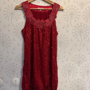 Röd glittrig ärmlös kortklänning - Supersnygg röd ärmlös klänning med glittriga paljetter och broderade detaljer vid halsringningen. Klänningen är kort och har en lös passform, perfekt för dig som vill sticka ut. Materialet känns lätt och festligt, och den breda axelremmen ger en cool look.