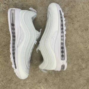 Nike Air Max 97 Triple White sneakers - Säljer ett par Nike Air Max 97 Triple White sneakers med ikonisk vågig design och synlig Air-enhet i sulan. Skorna är helvita med mesh och syntetiska paneler, snörning och diskret Swoosh-logga på sidan och plösen. Perfekta för dig som gillar clean och sportig stil.