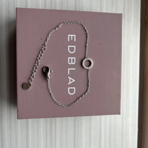 Silverarmband med cirkel från Edblad - Stilrent silverfärgat armband från Edblad med tunn kedja och en rund cirkel i mitten. Ena sidan av cirkeln är dekorerad med små glittrande stenar, medan den andra är slät. Mycket bra skick och använd fåtals gånger