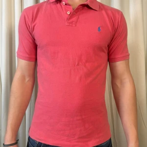 Rosa pikétröja från Polo Ralph Lauren - Snygg rosa pikétröja från Polo Ralph Lauren med klassisk krage och två knappar framtill. Tröjan har korta ärmar och den ikoniska blå logon broderad på bröstet. Tillverkad i mjuk bomull som är skön mot huden och har en slim passform.