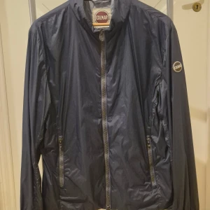 Mörkblå vindjacka från Colmar L - Snygg mörkblå vindjacka från Colmar Originals med dragkedja framtill och två fickor med dragkedja. Jackan har en stilren design med Colmar-logga på ärmen och hög krage. Perfekt för vår och höst när du vill ha något lätt och smidigt.