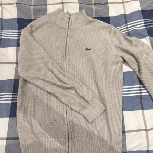 Beige kofta från Lacoste med dragkedja - Snygg beige kofta från Lacoste med ribbad struktur och hel dragkedja framtill. Klassisk ståkrage och det ikoniska gröna krokodilmärket på bröstet. Perfekt för dig som gillar stilrena och enkla plagg med premiumkänsla.