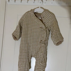 Newbie - overall, storlek 56 - Newbie overall storlek 56.  En beige och vit rutig overall med huva från Newbie. Den har tryckknappar framtill och muddar vid ärmslut och benslut.   Skickar med frakt eller avhämtning i Växjö. Vid flera köp kan du som köpare välja samfrakt. Fraktpriset ingår inte i priset. 