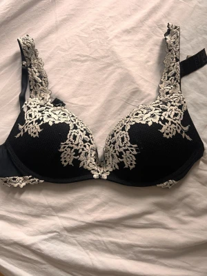 Svart intimissimi push-up bh med spetsdetaljer - Snygg svart push-up bh med vadderade kupor och dekorativa vita spetsdetaljer i blommigt mönster över kuporna och axelbanden. Bh:n har justerbara axelband och hakknäppning bak. Perfekt för dig som gillar en feminin och elegant stil.