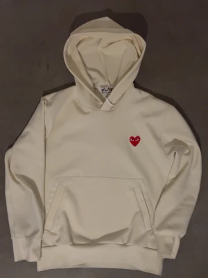 Vit hoodie från Comme des Garçons Play - Säljer en stilren off white hoodie från Comme des Garçons Play med det ikoniska röda hjärtat på bröstet. Hoodien är i bra skick men har ju såklart några små slitningar här och var (INGA HÅL). Inget som syns utan att man tänker på det🙌 Jag är 184 och väger 73kg och den sitter superbra på mig!✅ Nypris|2400kr, Mitt pris|600kr