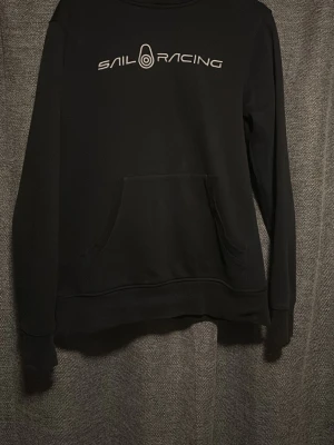 Svart hoodie från Sail Racing - Svart hoodie från Sail Racing med vit logga tryckt över bröstet. Klassisk känguruficka framtill och ribbade muddar vid ärmslut och nederkant. Perfekt för en avslappnad och sportig stil.