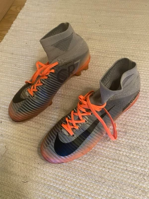 Nike Mercurial CR7 fotbollsskor - Snygga Nike Mercurial CR7 fotbollsskor med högt skaft och orangea snören. Skorna har en silvrig och svart ovandel med CR7-detalj på sidan och orange sula med dobbar. Perfekta för dig som vill sticka ut på planen. Storlek 39