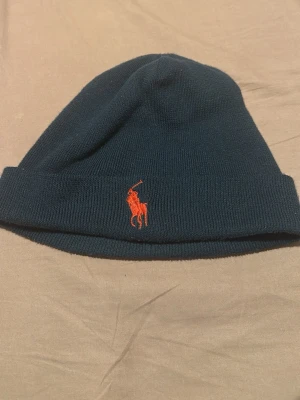 Mörkblå mössa från Polo Ralph Lauren - Snygg mörkblå stickad mössa från Polo Ralph Lauren med klassisk röd broderad logga framtill. Mössan har uppvikt kant och är tillverkad i mjukt stickat material som håller dig varm under kalla dagar.