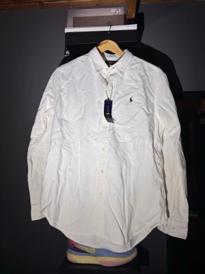 Vit skjorta från Ralph Lauren - Klassisk vit skjorta från Ralph Lauren med broderad logga på bröstet. Skjortan har lång ärm, knappar framtill och krage. Tillverkad i bomull och passar perfekt till en stilren look.