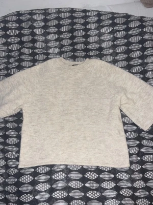 Beige stickad tröja med vida ärmar - En stilren beige stickad tröja med rund hals och vida, något kortare ärmar. Tröjan har en rak passform och är tillverkad i ett mjukt, melerat garn som ger en mysig känsla. Perfekt för lager-på-lager och enkel att matcha med olika outfits.