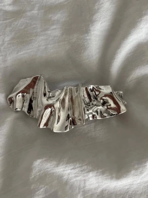 Silvrig metallisk hårklämma - Säljer en unik hårklämma i silvrig metall med skulptural, veckad design. Klämman har en blank, reflekterande yta och känns riktigt trendig och edgy. Perfekt accessoar för att lyfta din frisyr med en cool detalj.