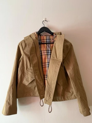 Beige trenchcoat med huva Burberry -   💓 Säljer en söt beige trenchcoat från burberry stilenmed klassiskt rutigt foder och huva, dragsko i midjan och långa ärmar. 😙 . Oops ”alla andra annonser med samma bild som min är felaktiga och säljer inte min jacka”