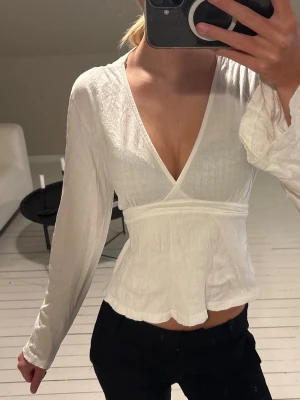 Vit topp/blus - Säljer en vit omlottblus med djup v-ringning och långa ärmar. Blusen har ett knytband i midjan som ger en snygg siluett och lätt volang nedtill. 