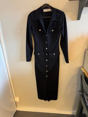 Svart långklänning Jazz Military - Stilren svart långklänning från Jazz Military med långa ärmar, markerad krage och knappar längs hela framsidan. Två bröstfickor med lock och dekorativa axelklaffar ger en cool touch. Perfekt för dig som gillar klassisk men edgy stil.