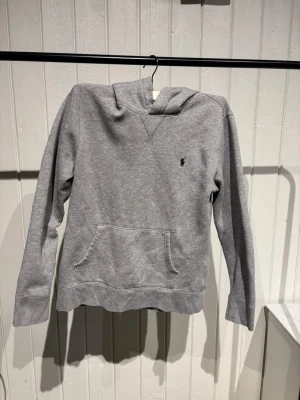 Grå hoodie från Polo Ralph Lauren - Klassisk grå hoodie från Polo Ralph Lauren med broderad logga på bröstet. Hoodien har huva, känguruficka och långa ärmar. Tillverkad i mjukt bomullsmaterial som är perfekt för en avslappnad stil. Passar folk som är runt 158