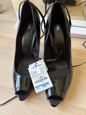 Svarta lackpumps från ZARA strlk 39 - Säljer ett par glansiga svarta pumps från ZARA i storlek 39. Skorna har öppen tå, smal klack och tunna ankelband som knyts runt vristen. Klacken är relativt hög, skulle vara ett par dansskor så den är ca 7-8 cm! Men verkligen supersnygga och faktiskt rätt bekväma. 