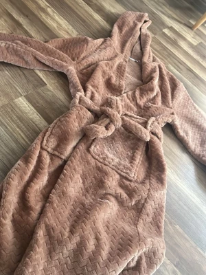 fluffig morgonrock - Supermjuk och mysig brun morgonrock från Name It med huva, fickor och knytskärp i midjan. Tillverkad i fluffigt, mönstrat material som känns extra skönt mot huden. Perfekt för lata morgnar eller kvällar hemma.