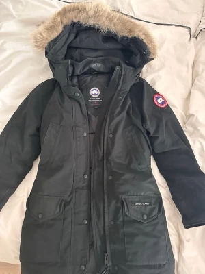 Svart jacka från Canada Goose - Säljer en svart jacka från Canada Goose med avtagbar huva och snygg pälskant. Jackan har två stora fickor framtill, dragkedja och knappar samt den klassiska Canada Goose-loggan på ärmen. Materialet är slitstarkt och jackan är fodrad för extra värme. Köpte den för 16000, priset kan diskuteras. Passar s och xs.