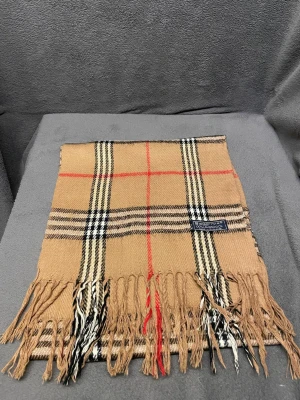 Burberry halsduk - Klassisk beige halsduk från Burberry med ikoniskt rutmönster i svart, vitt och rött. Halsduken har fransar i båda ändar och är tillverkad i mjukt material som känns lyxigt och stilrent. Perfekt accessoar för att lyfta din outfit. Halsduken är aldrig använd och nypris ligger runt 5000.            Skriv vid frågor, pris kan diskuteras 🤝