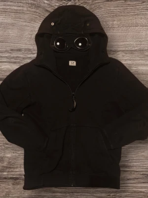 Äkta C.P. Company Hoodie - Svart hoodie från C.P. Company med dragkedja och ikoniska goggles i huvan. Tröjan har två fickor framtill och ribbade muddar. Perfekt för dig som vill sticka ut med en unik streetwear-look. Materialet känns mjukt och bekvämt.