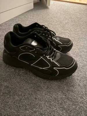 Dior b30 - Snygga svarta sneakers från dior med vita kontrastdetaljer och logga på hälen. Skorna har en chunky sula, meshpartier för extra andningsförmåga och klassisk snörning. Perfekta för dig som gillar en sportig och modern stil.