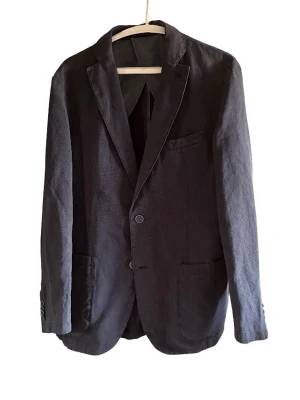 Svart klassisk kavaj med två knappar - Stilren svart kavaj med två knappar framtill, klassiska slag och tre fickor. Kavajen har en rak passform och är tillverkad i ett tyg som ser ut att vara bomull eller bomullsblandning. Perfekt för en dressad look.