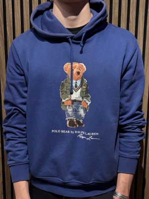Blå hoodie Polo Bear Ralph Lauren - Blå hoodie från Ralph Lauren med Polo Bear-tryck på bröstet. Klassisk huva med dragsko och ribbade muddar. Mjuk bomullsblandning och avslappnad passform. Perfekt för dig som gillar streetstyle och ikoniska prints.