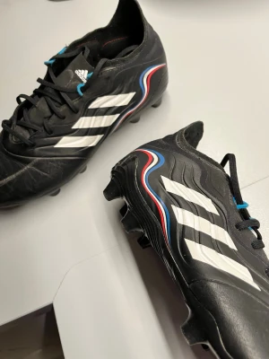 Svarta Adidas fotbollsskor - Säljer ett par svarta fotbollsskor från Adidas med vita ränder på sidorna och detaljer i blått och rött. Skorna har klassisk snörning och är tillverkade i syntetmaterial med en låg profil och dobbar för bra grepp på planen. Perfekta för dig som vill ha stilrena och funktionella skor.