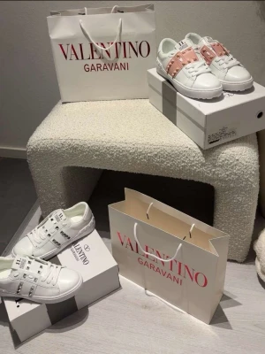Valentino Rockstud vita sneakers - Snygga vita sneakers från Valentino Garavani med ikoniska silverfärgade nitar och detaljer i läder. Modellen är låg och har klassiska snörningar. Finns även i en variant med ljusrosa band och nitar. Perfekta för dig som vill sticka ut med exklusiv design.