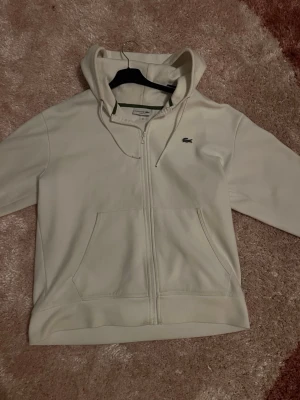 Vit hoodie med dragkedja från Lacoste - Snygg vit hoodie från Lacoste i klassisk passform. Hoodien har dragkedja framtill, två stora fickor och justerbar huva med snören. Ikoniska Lacoste-loggan i grönt broderad på bröstet. Perfekt för en clean och stilren look.