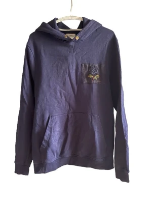 Mörkblå hoodie med broderad ficka - Snygg mörkblå hoodie med huva och känguruficka. På bröstet finns en liten ficka med broderad detalj och två knappar vid halsen. Tillverkad i mjukt bomullsmaterial, perfekt för en avslappnad och cool stil.