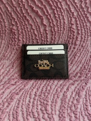 Svart kortfodral från Coach - Snyggt svart kortfodral från Coach med guldfärgad logga framtill. Fodralet har flera fack för kreditkort och är tillverkat i ett tåligt lädermaterial. Perfekt för dig som vill ha koll på korten med stil.