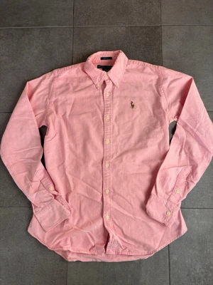 Rosa skjorta från Ralph Lauren - Klassisk rosa skjorta från Ralph Lauren med broderad logga på bröstet. Skjortan har lång ärm, knappar framtill och button-down krage. Slim fit-modell i mjuk bomull som ger en clean och stilren look.