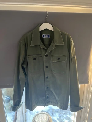 Olivgrön overshirt från Jack & Jones - Stilren olivgrön overshirt från Jack & Jones med klassisk krage, två bröstfickor med lock och knappar samt knappstängning framtill. Tillverkad i ett mjukt och slitstarkt material, perfekt för lager-på-lager-stil.