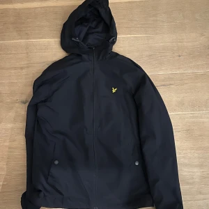  Lyle & Scott jacka - Snygg mörkblå vindjacka från Lyle & Scott med gul logga på bröstet. Jackan har huva, dragkedja framtill och två fickor med knappstängning. Tillverkad i ett lätt och tåligt material som passar perfekt för blåsiga dagar.