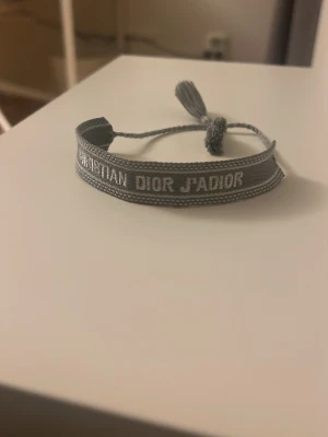 Grått tygarmband från Dior - Snyggt grått tygarmband från Dior med texten 'CHRISTIAN DIOR J'ADIOR' broderad i vitt. Armbandet har justerbar knytning med tofsar i ändarna för en cool och avslappnad look. Perfekt accessoar för att lyfta din outfit.
