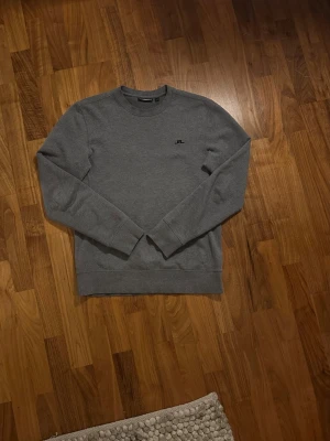 Grå sweatshirt från J.Lindeberg - Snygg grå sweatshirt från J.Lindeberg med rund halsringning och diskret svart logga broderad på bröstet. Tröjan har en klassisk passform och är tillverkad i mjukt material, perfekt för en avslappnad stil.