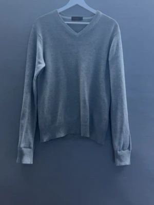 Grå stickad tröja (Morris) - En enkel och stilren ljusgrå stickad tröja från Morris med V-ringad hals. Tröjan har långa ärmar och ribbade muddar vid ärmslut och nederkant. Perfekt basplagg till garderoben och lätt att matcha med olika stilar.