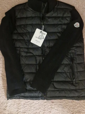 Svart cardigan från Moncler - Säljer en svart cardigan från Moncler med stickade ärmar och quiltad front. Jackan har dragkedja framtill, två dragkedjefickor och Moncler-logga på ärmen. Materialet är en mix av dunfylld nylon och stickad ull. Perfekt för kalla vinterdagar.