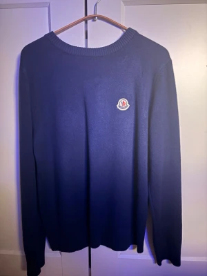 Mörkblå stickad tröja från Moncler - Mörkblå stickad tröja från Moncler med rund halsringning och Moncler-logga på bröstet. Tröjan har långa ärmar och ribbstickade muddar vid hals, ärmslut och nederkant. Perfekt för lager-på-lager och stilren look.