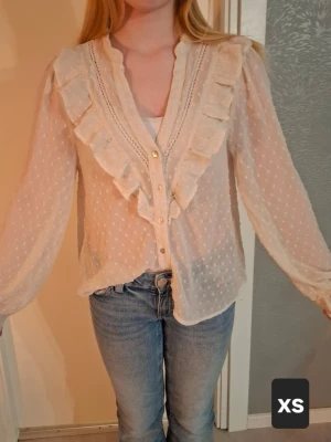 Vit volangblus med prickigt mönster - Söt vit blus med volangdetaljer längs knäppningen och V-ringning. Blusen har ett diskret prickigt mönster i tyget och är tillverkad i ett tunt, transparent material. Långa ärmar med lätt puff och knappar framtill ger en romantisk och trendig look.