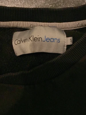 Svart sweatshirt Calvin Klein Jeans - Svart sweatshirt från Calvin Klein Jeans med stor silverfärgad logga och texten 'New York City EST. 1978' på bröstet. Tröjan har rund halsringning och ribbade muddar vid ärmslut och nederkant. Perfekt för en clean och stilren look.