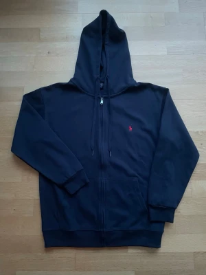Svart hoodie från Polo Ralph Lauren - Snygg svart hoodie från Polo Ralph Lauren med dragkedja, huva med dragsko och klassisk röd logga broderad på bröstet. Tillverkad i mjuk bomullsblandning och har två fickor framtill. Perfekt för en chill och stilren look.