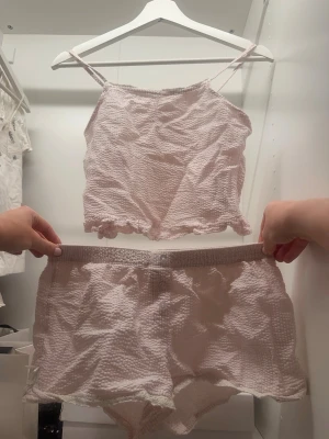 Ljusrosa loungewear-set med volang - Sött loungewear-set i ljusrosa med linne och shorts. Linnet har tunna axelband och volangkant nertill, medan shortsen har resår i midjan och matchande volangkant. Materialet är mjukt och strukturerat, perfekt för avslappnade dagar hemma. Shortsen i stl 158/164 och toppen i stl 146/152