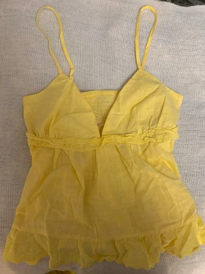 Gul linnetopp med spetsdetaljer och broderi  - Söt gul linnetopp med smala axelband och v-ringning. Toppen har en spetsliknande broderad detalj under bysten och volang längst ner. Perfekt för varma dagar och ger en fräsch sommarkänsla.