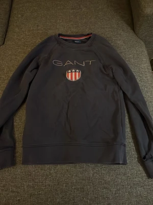 Mörkblå sweatshirt från GANT - Mörkblå sweatshirt från GANT med broderad logga och sköld med stjärnor och ränder på bröstet. Klassisk rund halsringning och ribbade muddar vid ärmslut och nederkant. Perfekt för en avslappnad och stilren look.