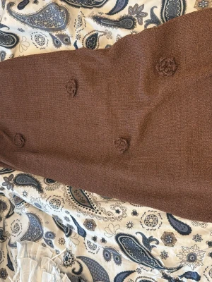 Brun stickad klänning  med blommor öppen rygg  - Mysig brun stickad klänning  med dekorativa stickade blommor. Kjolen har en rak passform och är perfekt för dig som gillar en avslappnad men ändå stilren look.