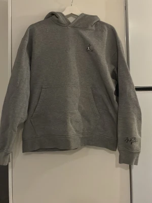 Grå Jordan hoodie med brodyr - Snygg grå hoodie från Jordan med klassisk huva och stor magficka. På bröstet sitter den ikoniska Jumpman-loggan broderad i vitt och på ärmen finns en diskret signaturdetalj. Hoodien är i mjukt material, perfekt för chill eller streetwear.